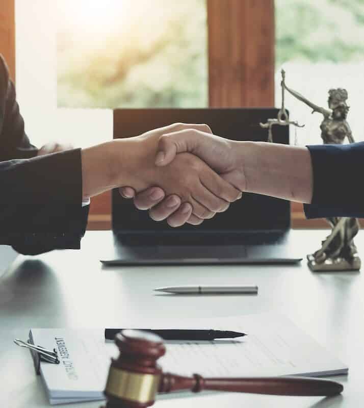 A legal professional and client shaking hands over a contract, with a gavel and a Lady Justice statue on the desk in a bright office.